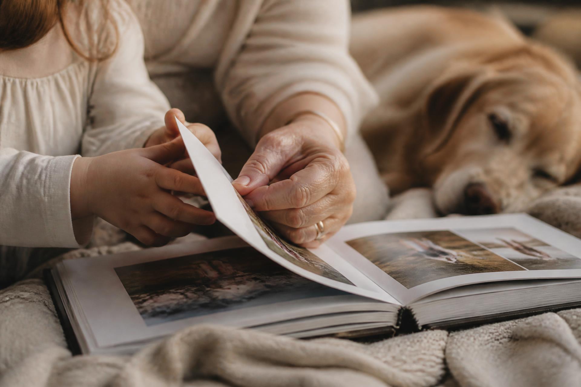 A grandmother and a young child turning the pages of a linen photo book, an older dog resting beside them.